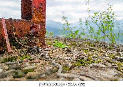 Old Rusty Screw Bolt At The Bottom Of An Abandoned Exposed Iron Construction Built On Weathered Concrete Base With Green Growth And Mountains In The Background