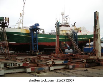 An Old And Rusty Ship In The Dry Dock Is Being Repaired And Repainted. A Person In Yellow Oil Rig Is Working On It.