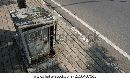 Old, rusty utility box on a city sidewalk beside an asphalt road, showing signs of decay and urban neglect in the midday sunlight.