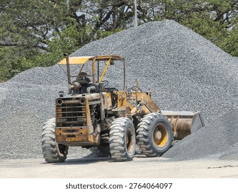 An old, rusty wheel loader parked in front of large piles of crushed gravel at a construction site.