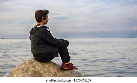 Old Sad Depressed Woman Sit Alone On Rocks, Looking At Distant Sea / Seascape Horizon. Time To Go, Say Goodbye. Miss Someone. Desire, Hope To Go Far Away. Unhappy Woman Feel Lonely At Seaside Outdoor