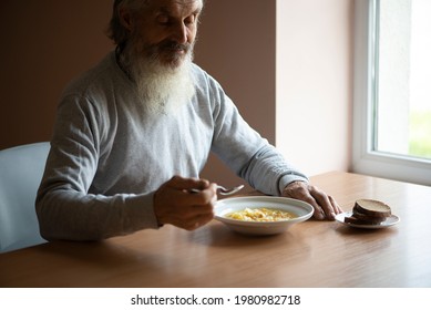 Old Sad Man With A Long Gray Beard Sitting By The Table And Eating Soup And Bread