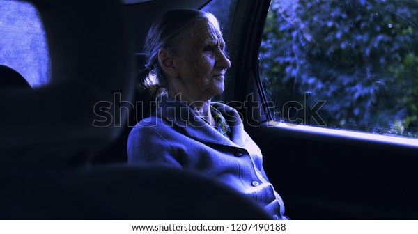 An old sad woman with deep\
wrinkles rides in the car at night. Grandmother sits in the\
car.
