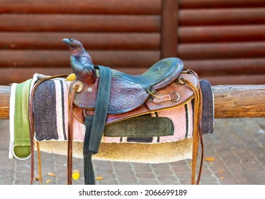 Old saddle on fence against the wooden wall. Sport leather saddle, harness for horses on daylight. Western horse saddles on a rack,  ready for dressage training. Equestrian sport background, vintage. 