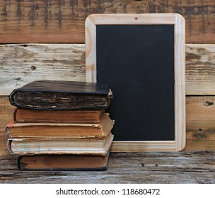 old school books are stacked on a desk in front of a blackboard.