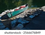 Old scratched green canoe framed between stones on edge of water, Rope hanging down and life jackets inside. Reflections of sky and clouds in calm smooth blue and green water. On a sunny day no people