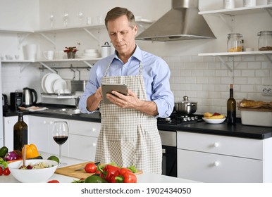 Old senior 50s man wearing apron using tablet preparing vegetable salad in kitchen. Middle aged chef searching recipes, ordering food online, watching cooking videos preparing healthy food at home. - Powered by Shutterstock