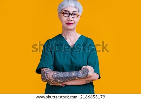 old senior asian female professional doctor grey hair and tattoo with glasses wear hospital uniform standing with confident and cheerful expression studio shot yellow background