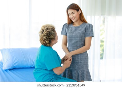 Old Senior Asian Happy Healthy Recovery Mother Patient In Hospital Uniform Sitting Smiling On Bed While Young Woman Daughter Visiting Standing Holding Hands With Love And Care In Clinic Wardroom.