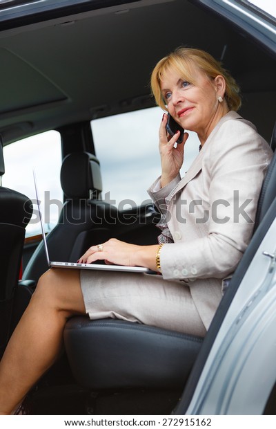 Old senior business woman 60-65 years talking by\
mobile cell phone in the back of a car and holding her Laptop\
Computer pc in lap