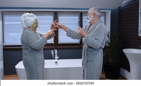 Old senior couple man and woman in bathrobe are dancing in bathroom having fun laughing enjoying leisure time and music. Elderly grandmother with facial green mask and grandfather with shaving foam - Powered by Shutterstock