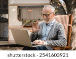 Old senior elderly man grandfather using laptop for remote work, having videocall conference online while caravanning by trailer camper van motor home