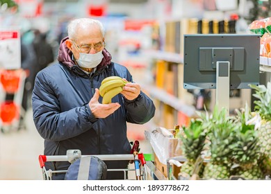Old senior european man wearing protective facial mask looking at banana in the supermarket. Shopping during COVID-19 concept