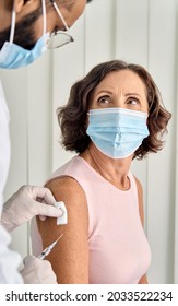 Old Senior Lady In Facemask Getting Covid19 Protective Vaccine Receiving Antiviral Injection In Arm Sitting In Clinic Hospital With Young Doctor In Surgery Mask. Vaccination Safety Concept. Vertical.