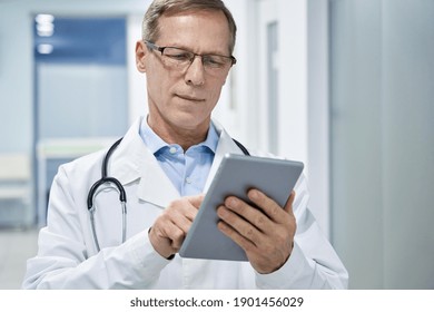 Old Senior Male Doctor Holding Digital Tablet In Hands Using E Health Technology Apps In Hospital Consulting Patients Online. Modern Tech Medical Healthcare Virtual Telehealth, Tele Medicine Concept.