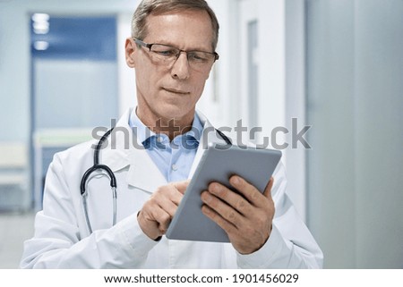 Old senior male doctor holding digital tablet in hands using e health technology apps in hospital consulting patients online. Modern tech medical healthcare virtual telehealth, tele medicine concept.