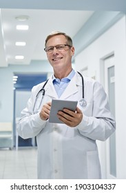 Old Senior Male Professional Doctor Physician Holding Digital Tablet Computer Looking At Camera In Hospital. Medical Healthcare Technology For Online Tele Medicine E Health Consultation Concept.