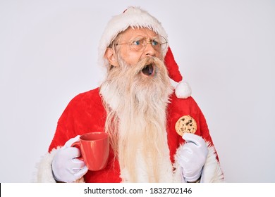 Old Senior Man With Grey Hair And Long Beard Wearing Santa Claus Costume Eating Cookies Angry And Mad Screaming Frustrated And Furious, Shouting With Anger. Rage And Aggressive Concept. 