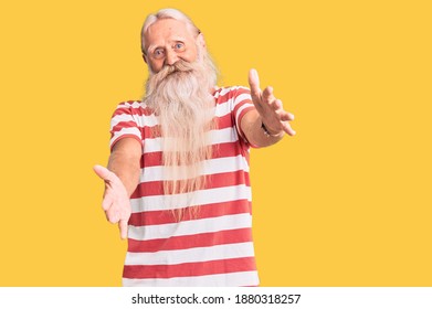 Old Senior Man With Grey Hair And Long Beard Wearing Striped Tshirt Looking At The Camera Smiling With Open Arms For Hug. Cheerful Expression Embracing Happiness. 