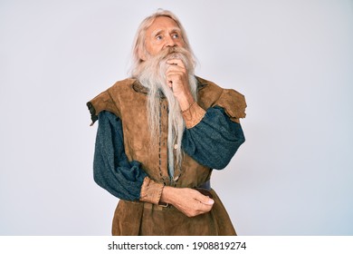 Old senior man with grey hair and long beard wearing viking traditional costume with hand on chin thinking about question, pensive expression. smiling and thoughtful face. doubt concept. 
