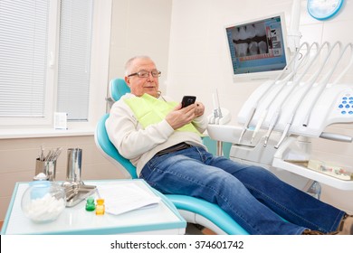 Old Senior Man Sitting In A Chair In Dental Clinic Read Or Write Sms On Cell Mobile Phone.