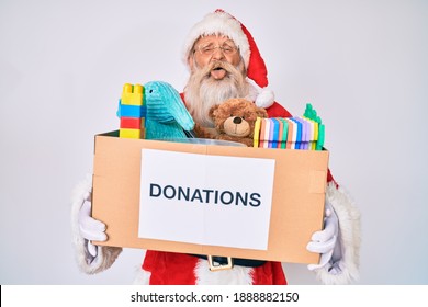 Old senior man wearing santa claus costume holding donation box sticking tongue out happy with funny expression. 