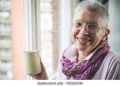 Old Senior Woman Drinking A Warm Beverage, Coffee Or Tea, Looking Through The Window