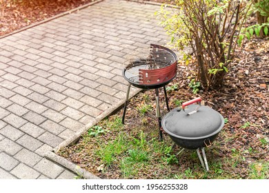 An Old Shabby Metal Barbecue Grill Stands In The Backyard On The Ground With Grass By The Bushes And Brick Path In Summer Day