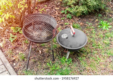 An Old Shabby Metal Barbecue Grill Stands In The Backyard On The Ground With Grass By The Bushes And Brick Path In Summer Day