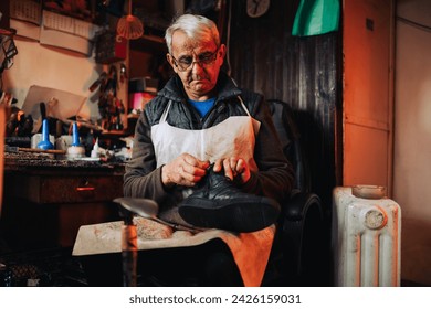 An old shoemaker is sitting at his cobbler workshop and examining boots. A senior cobbler is fixing boots at his shop. Small business owner is running a cobbler workshop. Craftsmanship concept. - Powered by Shutterstock