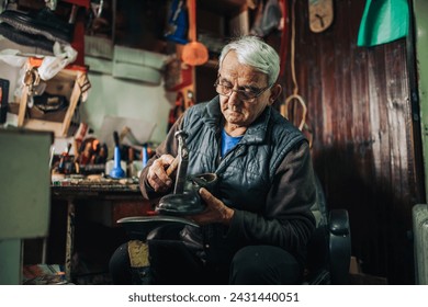 An old shoemaker is sitting in his small workshop and repairing shoes. An artisan is fixing footwear at his small shoemaker workshop and using his tool. Senior craftsman is repairing footwear at shop. - Powered by Shutterstock