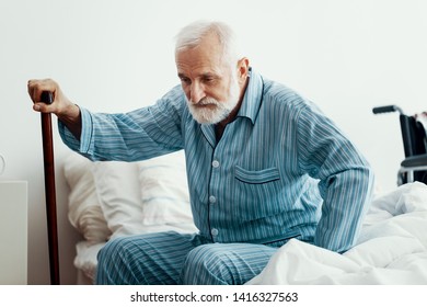 Old Sick Man With Grey Beard And Hair Wearing Blue Pajamas And Sitting On Bed At Home