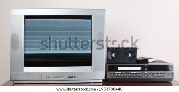 Old silver vintage TV with noise and interference\
on the screen and VCR on the background of the wallpaper. 1980s,\
1990s, 1970s.