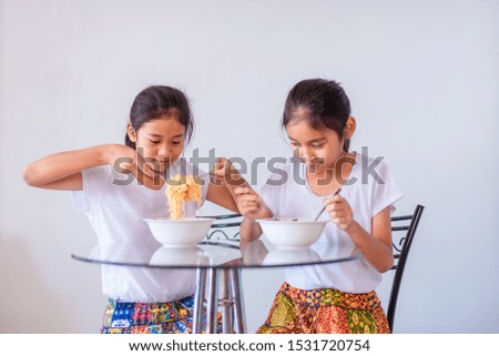 The old sister and the young sister are eating yellow noodles in the white bowls. It's delicious but spicy. Both of them wore a white T-shirt with a smiling face. The white background is behind them.
