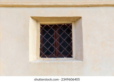 Old small square window closed with metal bars on wall of old building.