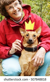 Old, Smiling Woman Holding Paper Crown On Stick Close To Dog's Muzzle. Dog In Funny, Fake Crown With Ownerin Park.