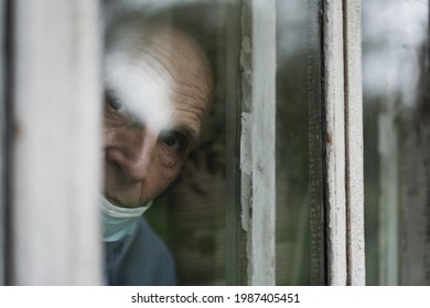 Old solitary man wearing face mask looking through window of nursing house.