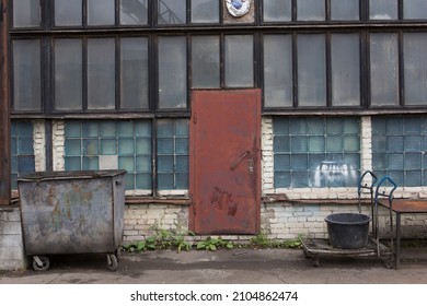 old soviet constructivist industrial building exterior facade with red steel door, large windows, blue glass bricks. Rusty, trolley, weathered garbage container in front