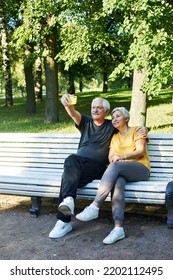 Old Spouses Make Selfie Picture Or Video Call Sit On Bench In Summer Park. Grey-haired Couple Look At Cell Phone Screen Make Videocall Outdoor Use Modern Tech, Enjoy Online Fun Spend Time Outside