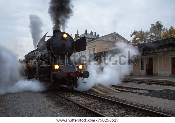 Old Steam train on Railway\
Station of Nova Gorica, Slovenia. Old Steam Locomotive of black\
color is driving and leaving a lot of smoke from chimney and\
vapour.