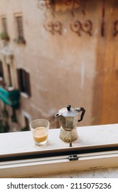 Old Steel Italian Geyser Coffee Maker On A Marble Windowsill With A Cup Of Hot Espresso With Milk. Coffee Prepared In A Geyser Coffee Maker Is Cooled In The Window. Morning Coffee Routine In Italy