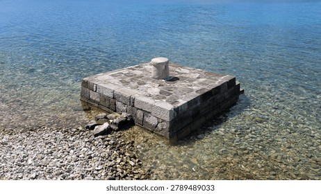 An old stone block structure with a central bollard stands in crystal clear shallow sea water next to a rocky pebble beach