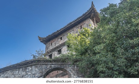 The old stone building has empty window openings. Curved roof eaves. A fragment of an arched bridge in the foreground. Branches of green trees. The blue sky. China. Fenghuang Ancient Town.  Phoenix.