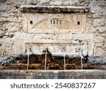 An old stone fountain dated 1877 with four water spouts in a rustic setting.