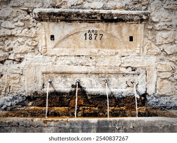 An old stone fountain dated 1877 with four water spouts in a rustic setting. - Powered by Shutterstock