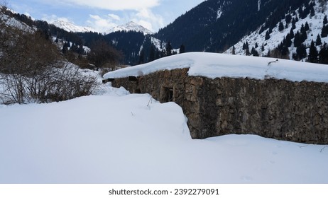 An old stone prison or hut in the mountains. Snowy mountains, forest and clouds against a blue sky. Tall dry bushes peek out from under the snow. Birch and spruce trees are visible in the distance. - Powered by Shutterstock