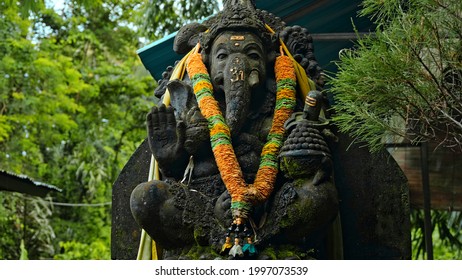 An old stone statue of Ganesha covered with moss with a wreath of yellow flowers on his neck standing on the street with green plants on the background - Powered by Shutterstock