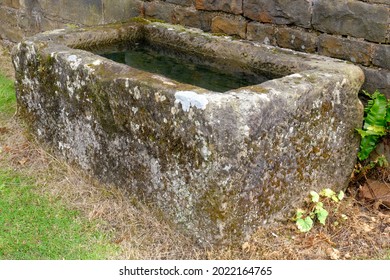An old stone trough stands by an old stone wall with coloured lichen, and moss emphasising age. - Shutterstock ID 2022164765