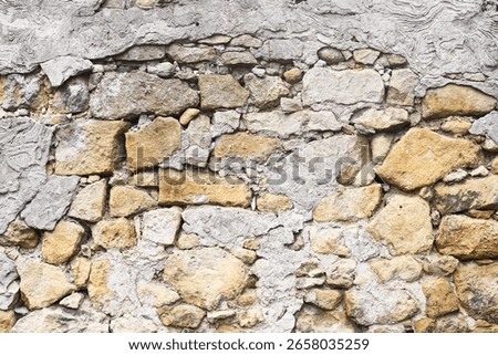Old stone wall background. Rough surface of mixed stones and cement. Natural gray and yellow texture. Rustic uneven construction detail. Weathered exterior of vintage traditional architecture.