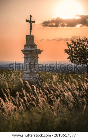An old stone wayside cross standing in a tall grass field during a dramatic orange sunset in the countryside.